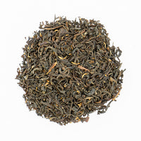 A circular pile of loose-leaf tea leaves, predominantly dark brown and black with lighter tan and yellowish-brown accents, photographed from directly above on a solid white background.