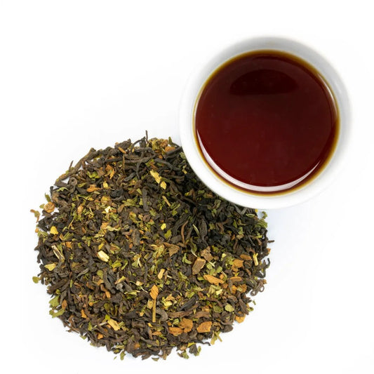 A pile of loose leaf Mint-Chilla Chai-Nilla Tea with green and brown leaves sits next to a white cup filled with dark brown tea on a white background.