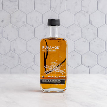 A bottle of Runamok Vanilla Bean Infused Maple Syrup with amber liquid, a black cap, and a white label, set on a white marble surface against a white hexagonal tile background.