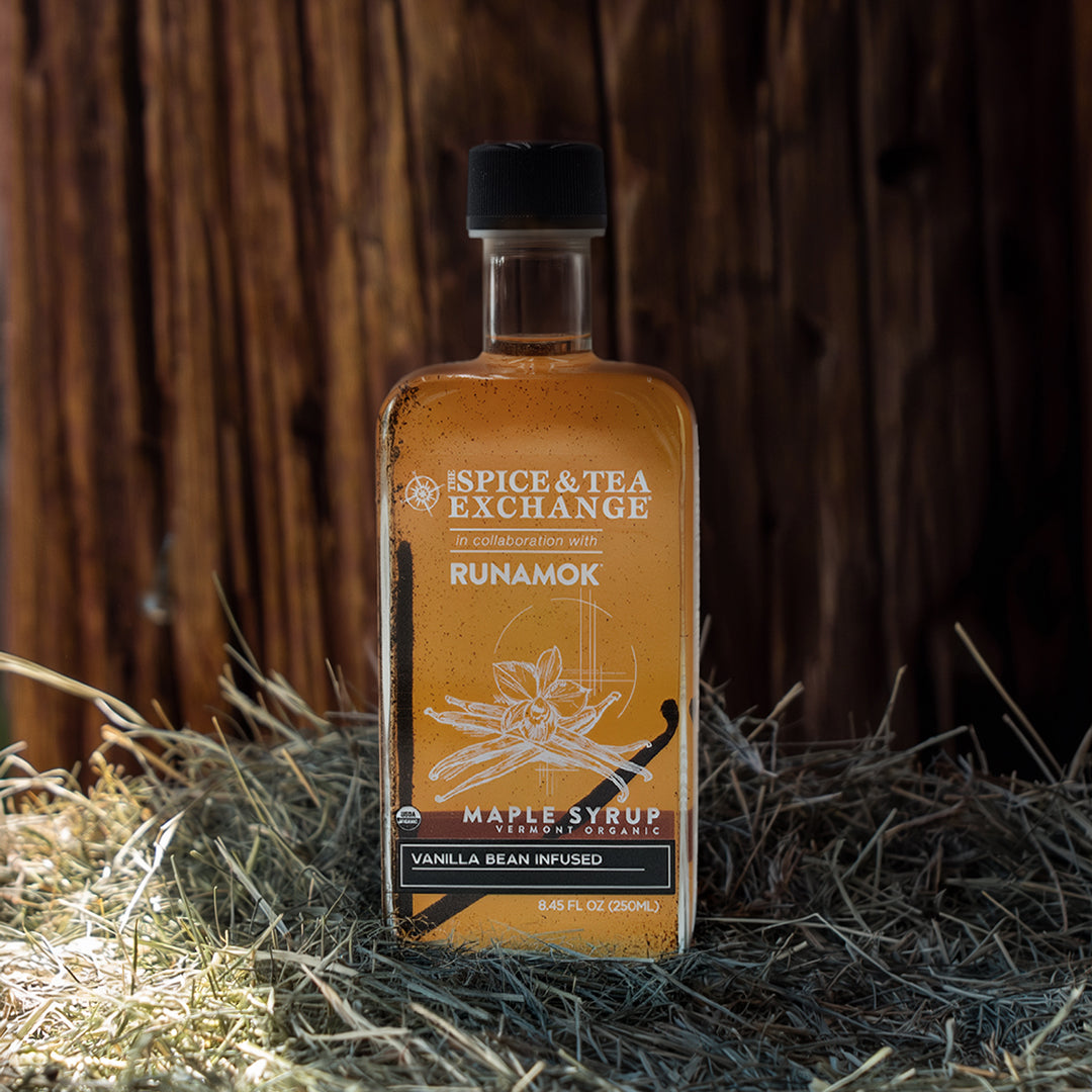 A bottle of Spice & Tea Exchange Runamok vanilla bean infused maple syrup, filled with amber liquid, set on a bed of straw against a dark wooden background.