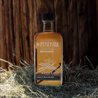 A bottle of Spice & Tea Exchange Runamok vanilla bean infused maple syrup, filled with amber liquid, set on a bed of straw against a dark wooden background.