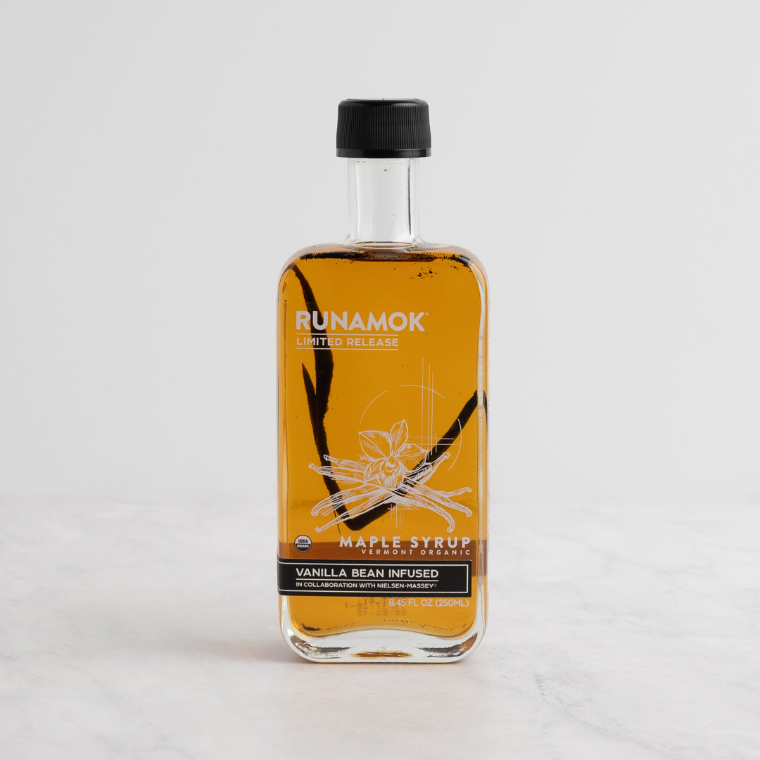 A bottle of Runamok Vanilla Bean Infused Maple Syrup with amber liquid, a black cap, and a white/black label, set on a light marble surface against a white background.