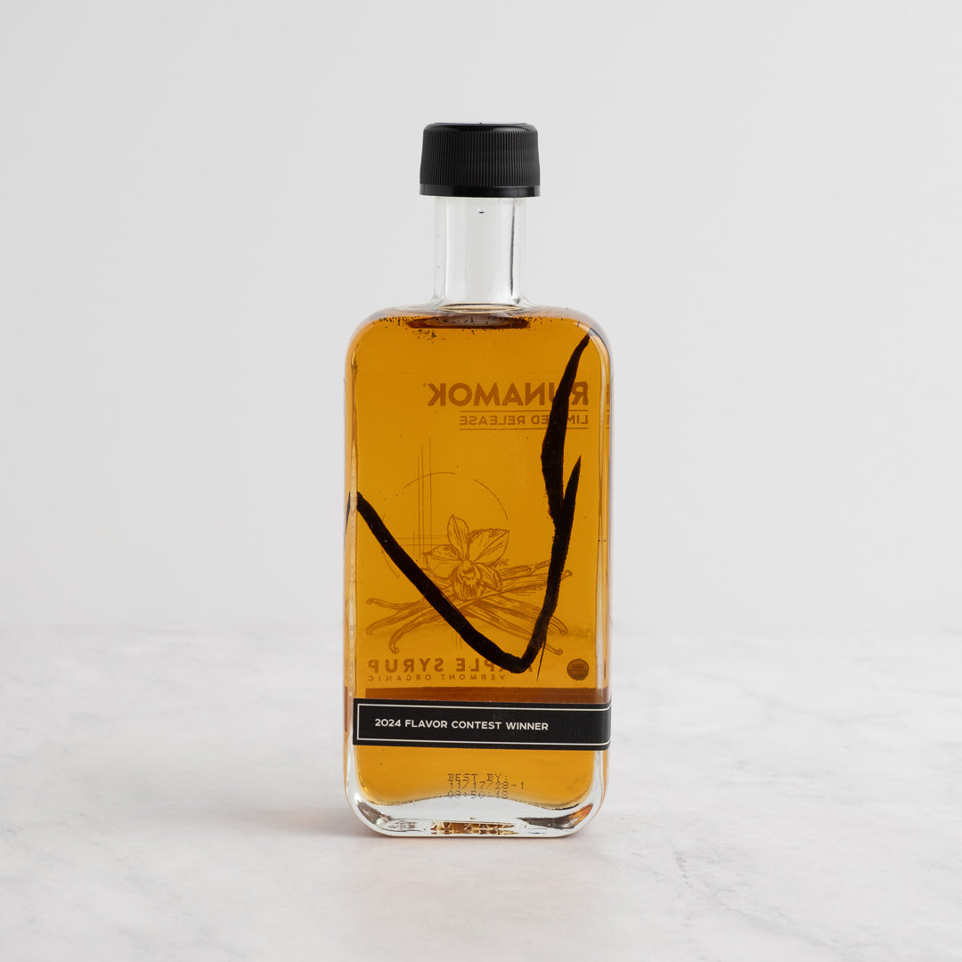 A bottle of Spice & Tea Exchange Runamok maple syrup with vanilla bean infusion, featuring golden-amber liquid inside a clear glass bottle with a black cap, set against a plain white background. Back side.