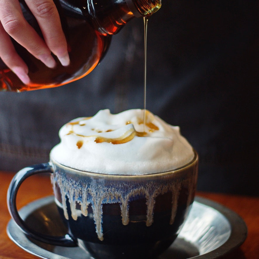 A close-up of a hand pouring amber syrup from a dark bottle into a frothy latte in a glossy dark ceramic mug with drips, set on a saucer against a dark background.