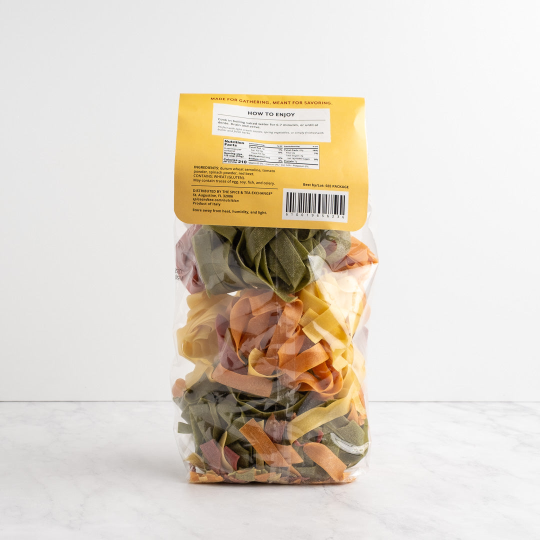 A clear plastic bag of mixed dried pasta sheets with a yellow label, standing upright on a light marble surface against a white background. Pappardelle-Primavera back label.