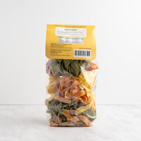 A clear plastic bag of mixed dried pasta sheets with a yellow label, standing upright on a light marble surface against a white background. Pappardelle-Primavera back label.