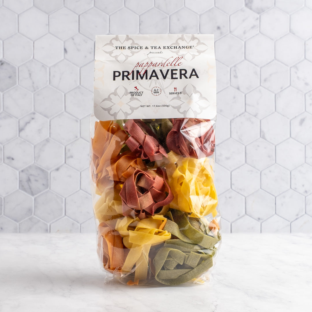 A bag of The Spice & Tea Exchange Pappardelle-Primavera pasta, featuring multicolored flower-shaped flat pasta in yellow, green, orange, and red, set against a white marble surface and hexagonal tile background.