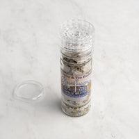 A clear spice grinder labeled Parfume de Sel Spice Blend from The Spice & Tea Exchange sits on a light marble surface, with its detached cap resting beside the bottle.