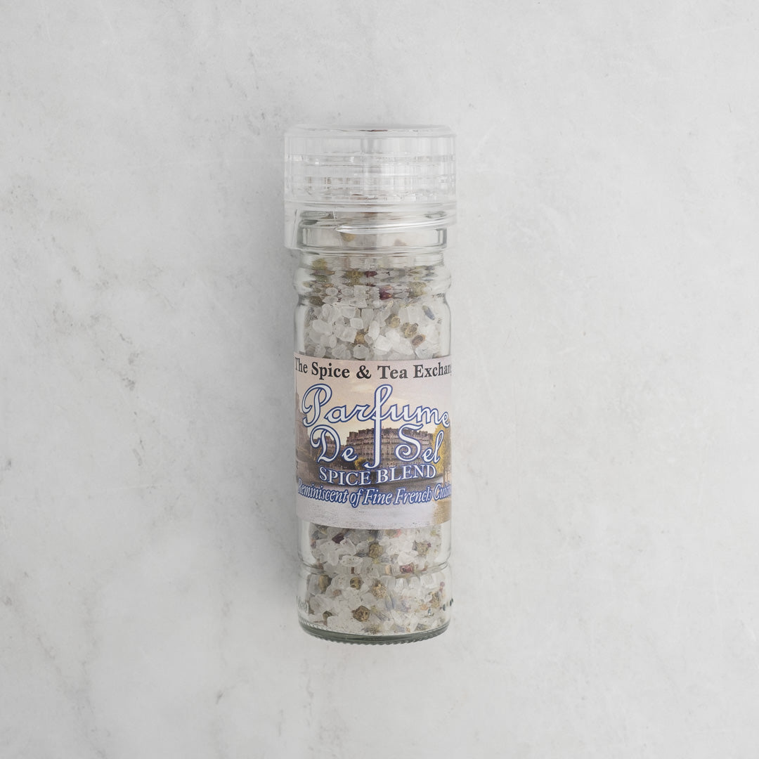 An upright, clear Parfume de Sel Spice Blend grinder bottle from The Spice & Tea Exchange, filled with French-inspired gourmet salt and spices, sits on a light marble surface.