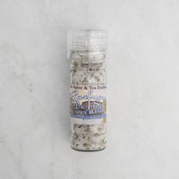 An upright, clear Parfume de Sel Spice Blend grinder bottle from The Spice & Tea Exchange, filled with French-inspired gourmet salt and spices, sits on a light marble surface.