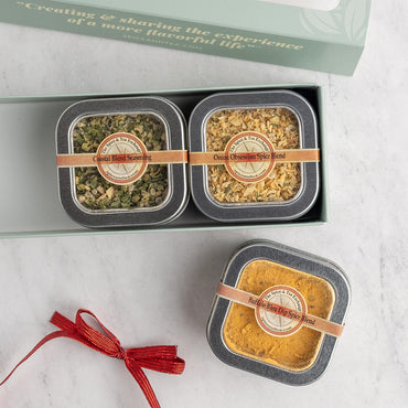 A gift set of three small round metal spice tins displayed on a light marble surface. The set includes two tins in an open mint green box labeled and one loose tin below filled with spice blends.