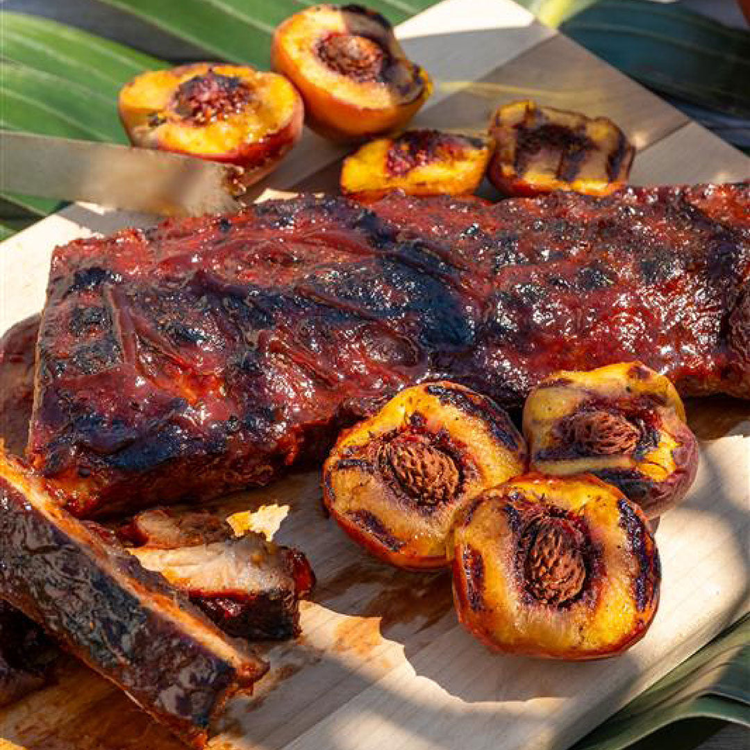 Grilled barbecue ribs on a wooden board, surrounded by halved, grilled peaches with char marks. The scene suggests a summer barbecue setting.