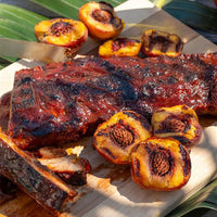 Grilled barbecue ribs on a wooden board, surrounded by halved, grilled peaches with char marks. The scene suggests a summer barbecue setting.