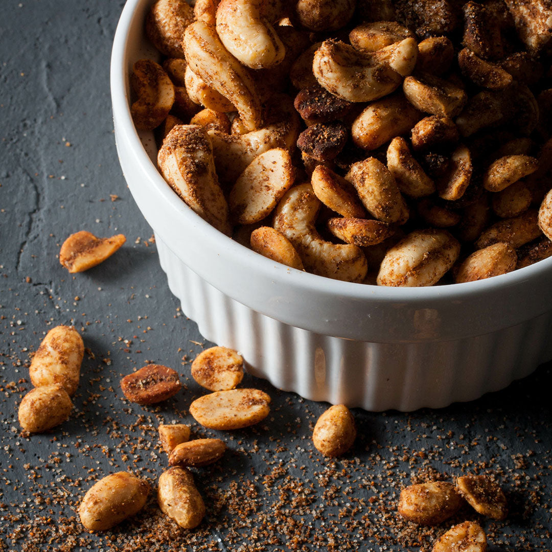 Crispy roasted cashews seasoned with spices, served in a white ribbed ceramic bowl with some scattered on a dark textured surface.