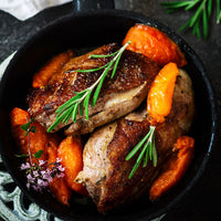Roasted chicken pieces in a black skillet with orange carrots, rosemary sprigs, and small purple flowers on a dark tabletop.