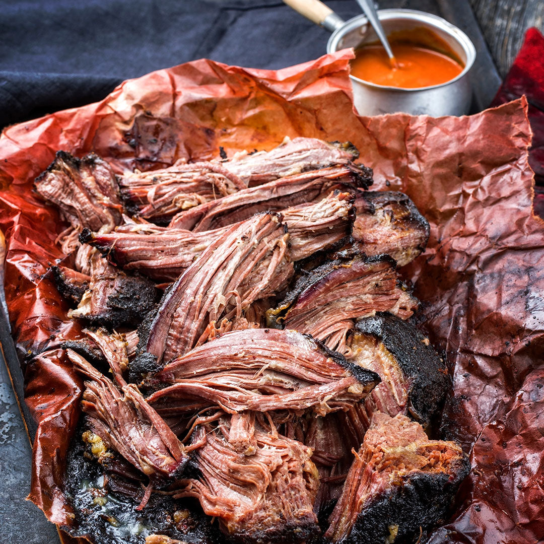 Shredded cooked meat on brown paper with a small metal saucepan of orange-red sauce in the background.