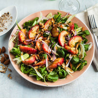 A top-down view of a fresh salad served on a peach-colored plate, featuring green leafy greens, sliced red fruit, red onion, and chopped nuts, set on a light gray surface with a fork and small side dishes.