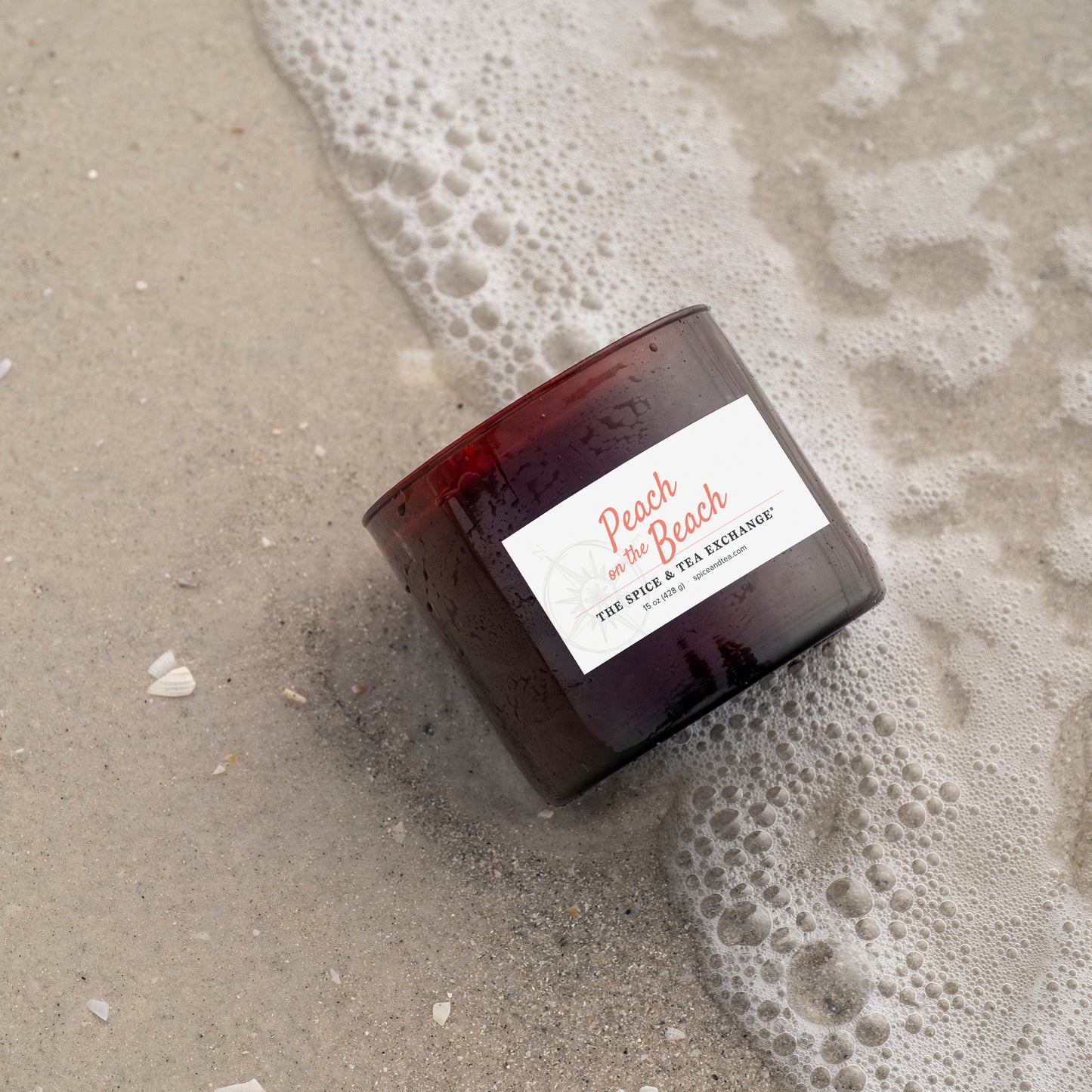 Candle labeled 'Peach on the Beach' on wet sand with foam