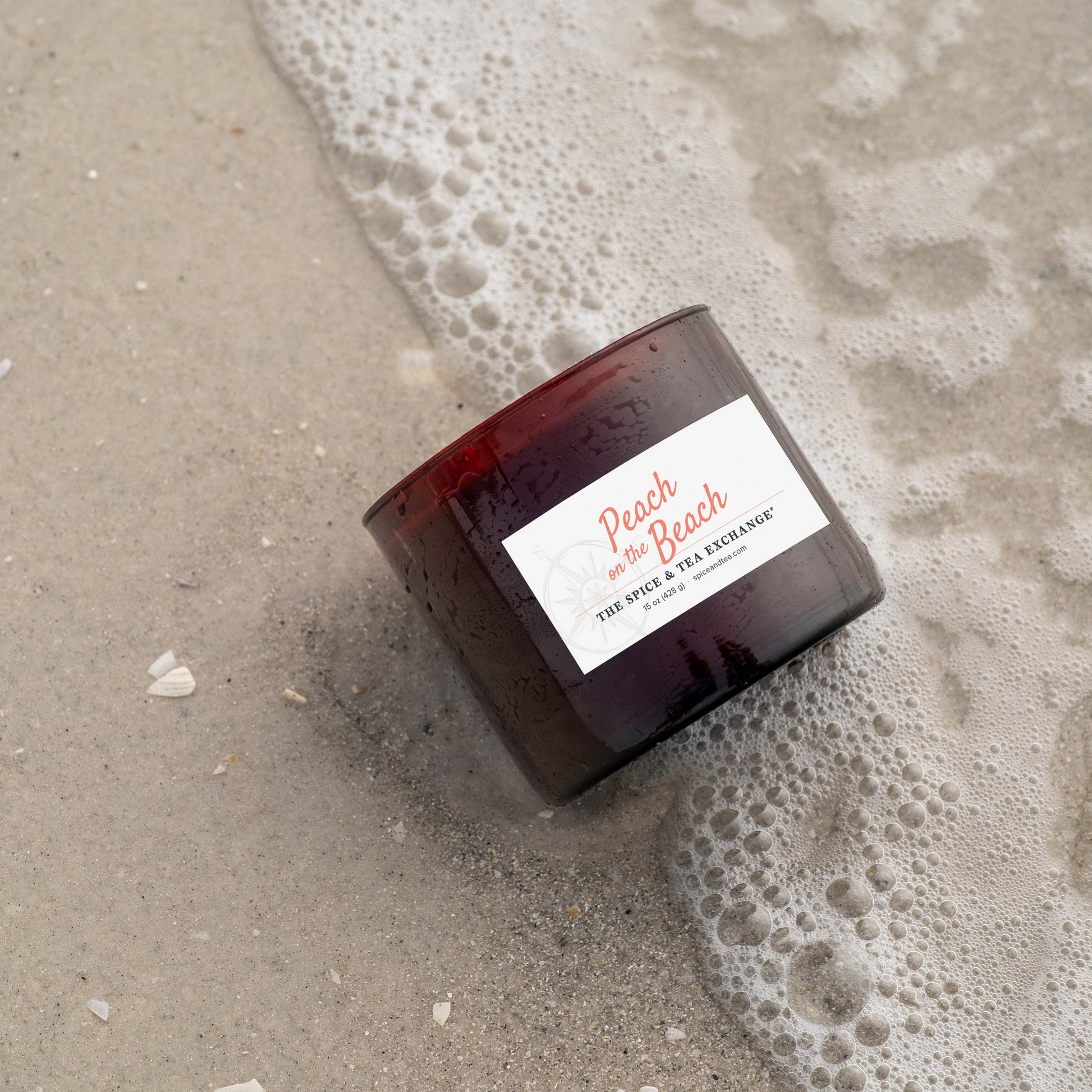 Candle labeled 'Peach on the Beach' on wet sand with foam