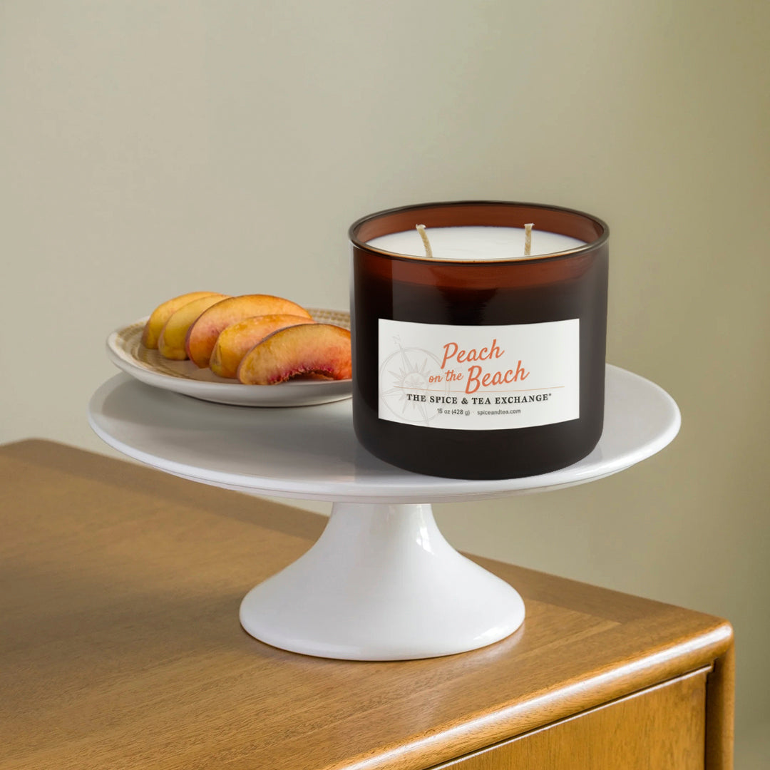 Candle labeled 'Peach on the Beach' by The Spice & Tea Exchange on a white pedestal with peaches in the background.