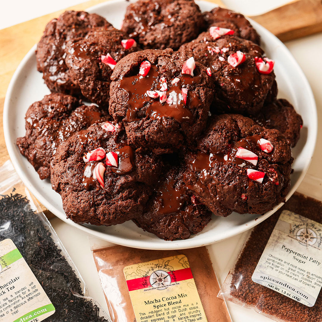 A white plate piled with chocolate chip cookies topped with drizzled chocolate and crushed candy canes, surrounded by ingredient bags including chocolate chips, mocha cocoa mix, and peppermint patty sugar on a wooden surface.