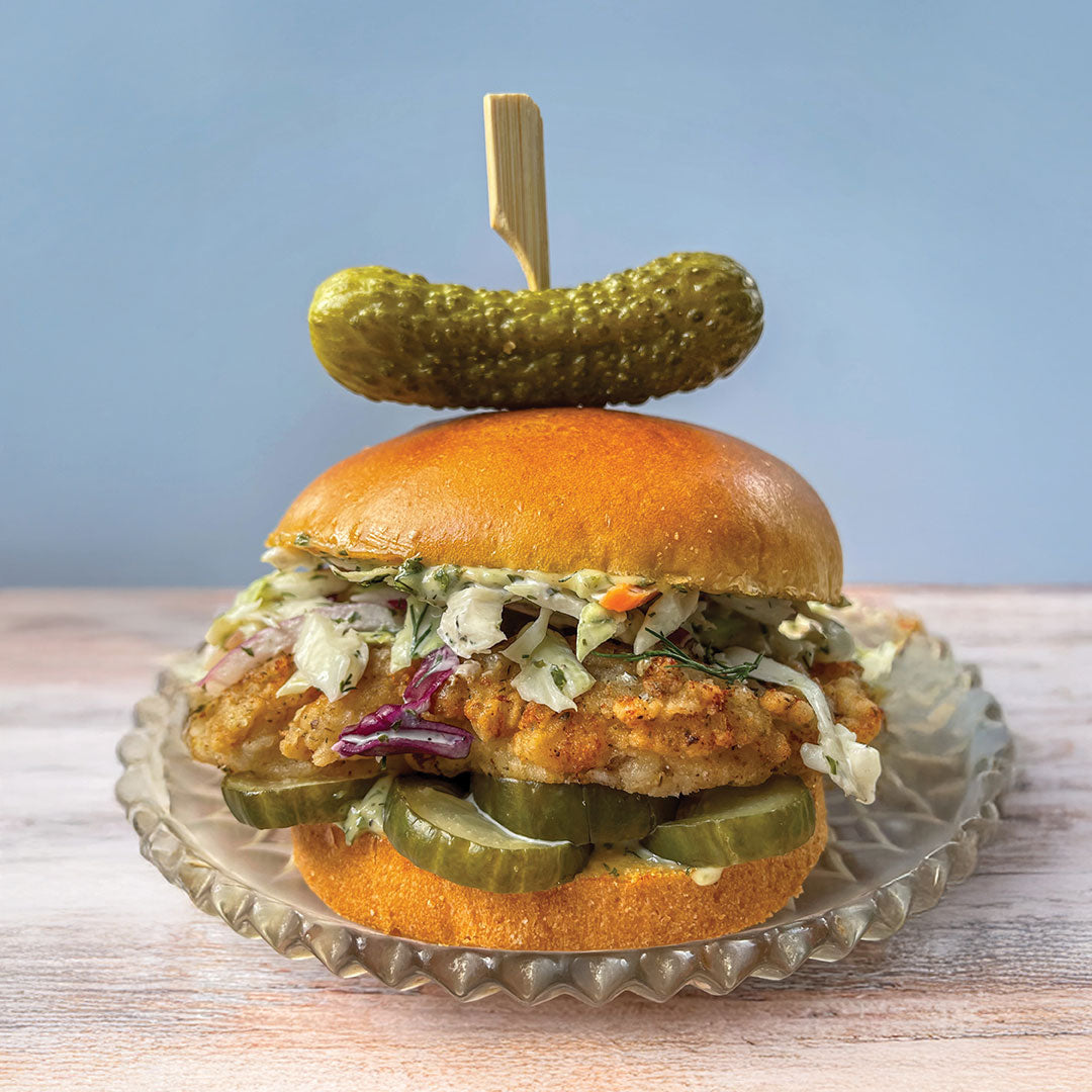 A fried chicken sandwich with coleslaw and pickle slices on a brioche bun, served on a glass plate. A whole pickle is skewered on top with a wooden pick. The background is light blue.