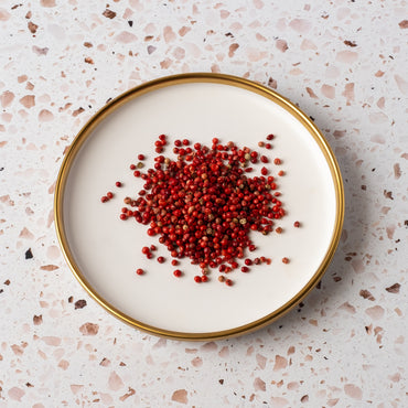 A round plate with a gold rim holds a pile of Pink Pepperberry, set on a pink and white terrazzo surface.