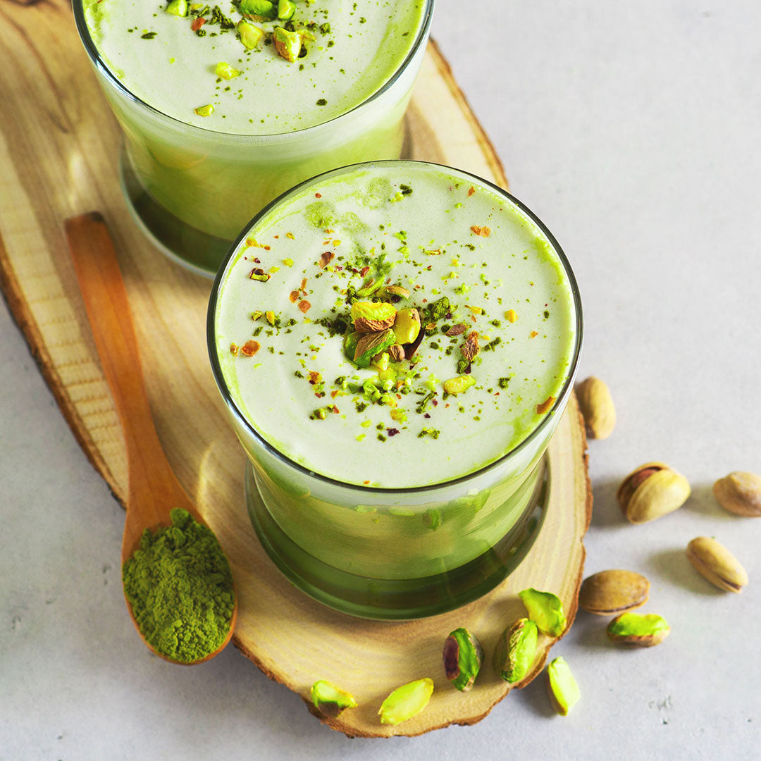Two clear glasses filled with a light green pistachio matcha drink, topped with chopped pistachios and a sprinkle of ground matcha, arranged on a wooden serving board with a wooden spoon of green powder and scattered pistachios on a light gray surface.