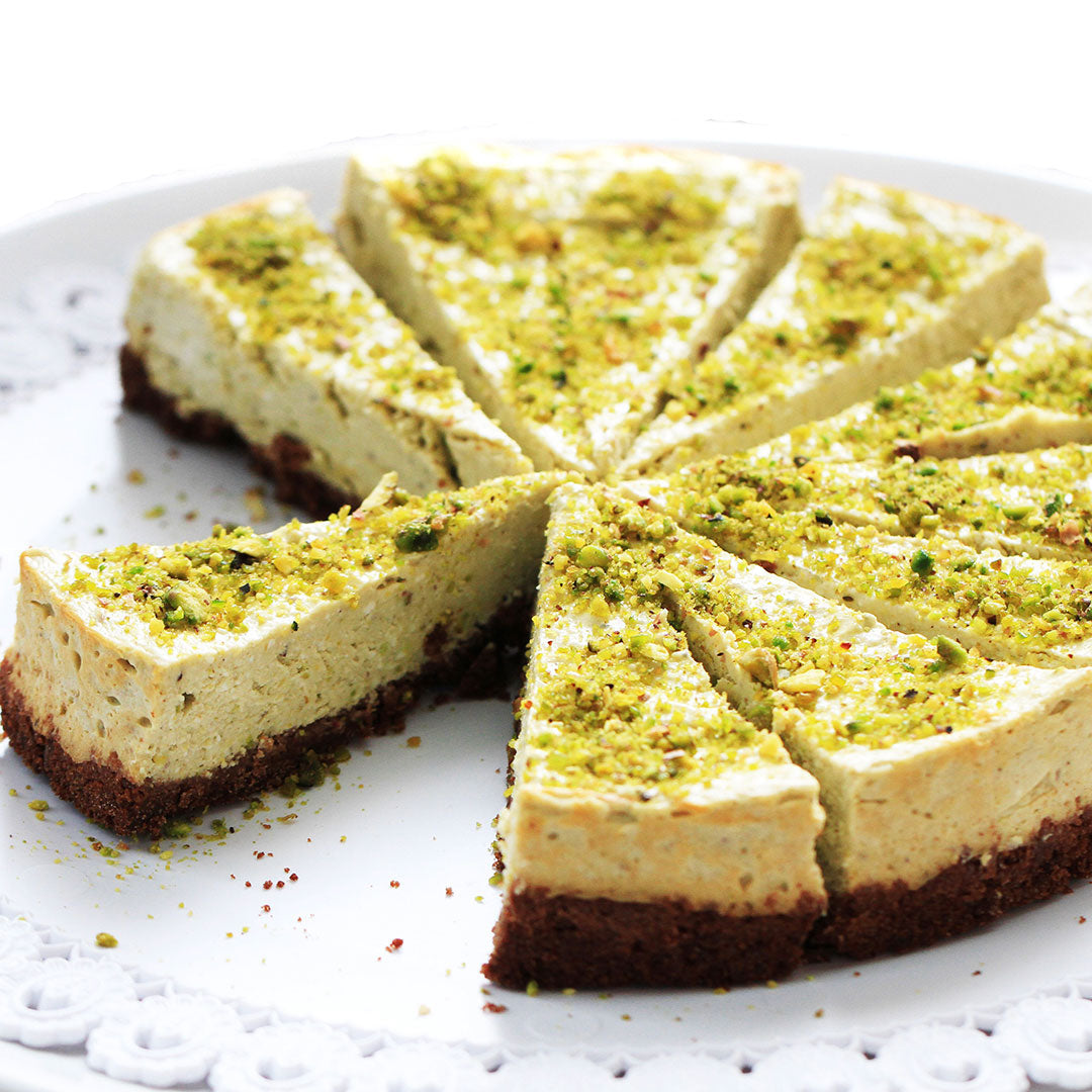 A sliced pistachio cheesecake with a dark chocolate cookie crust, topped with chopped green pistachios, served on a white plate.
