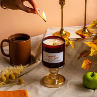A lit candle in a glass pedestal holder on a draped fabric surface, with a hand holding a match near the flame. A brown mug, gold candlesticks, yellow leaves, and a green apple surround it in a styled autumn scene.