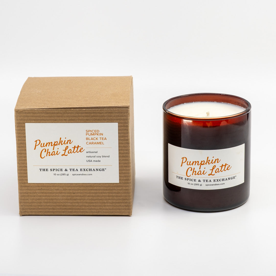 A Pumpkin Châi Latte scented candle in an amber glass jar next to its brown kraft cardboard box, both featuring white labels with orange text, on a clean white background.