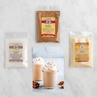 Three packaged baking ingredients arranged on a light marble surface: a clear bag of pie flour, a brown bag labeled pumpkin pie spice blend, and a clear bag of sugar. A small printed photo in the center shows pumpkin pie malt drinks with whipped topping and cinnamon.
