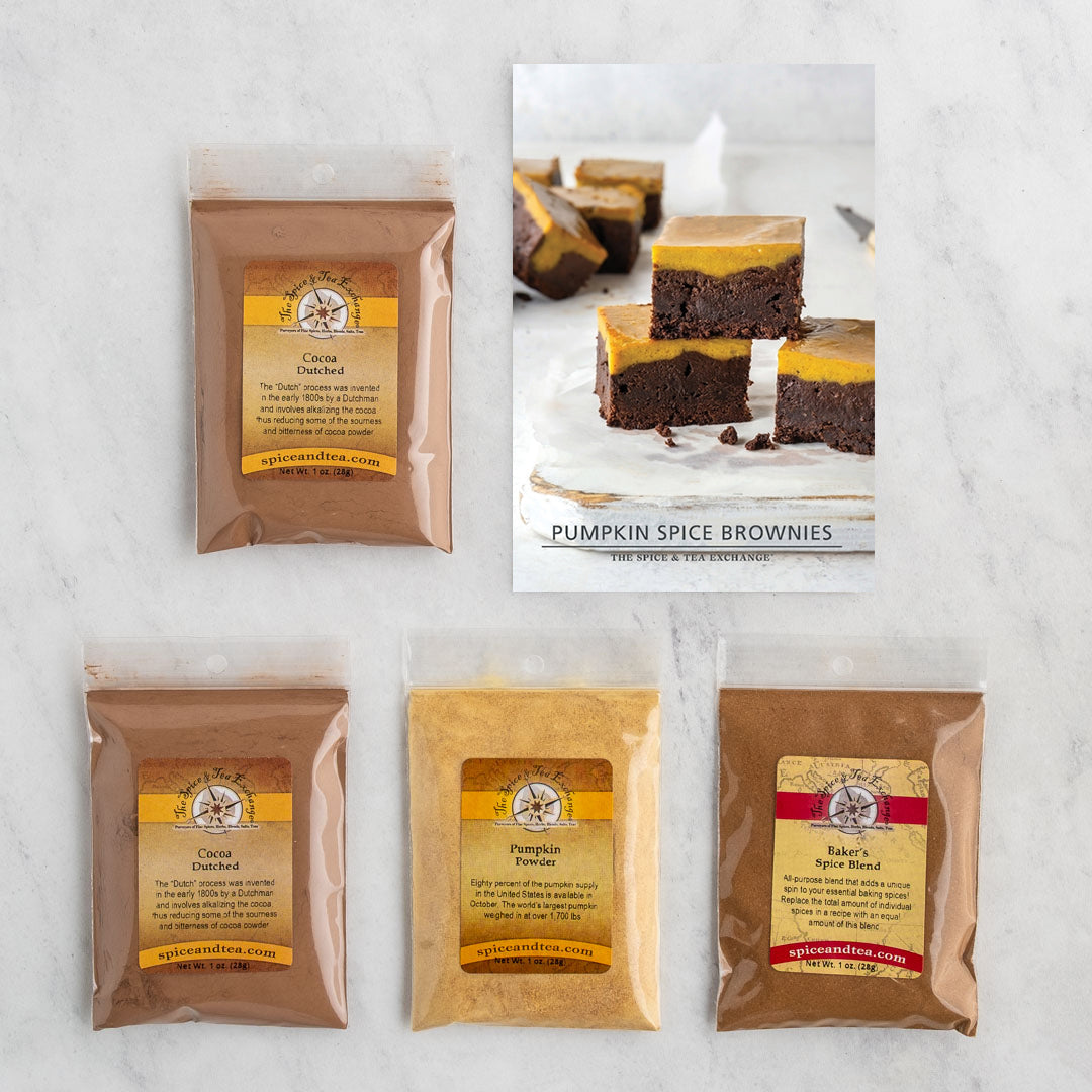 A flat-lay of four packaged pumpkin spice brownie mix ingredients on a light marble surface, alongside a printed recipe card showing finished pumpkin spice brownie bars.