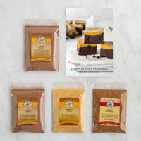 A flat-lay of four packaged pumpkin spice brownie mix ingredients on a light marble surface, alongside a printed recipe card showing finished pumpkin spice brownie bars.