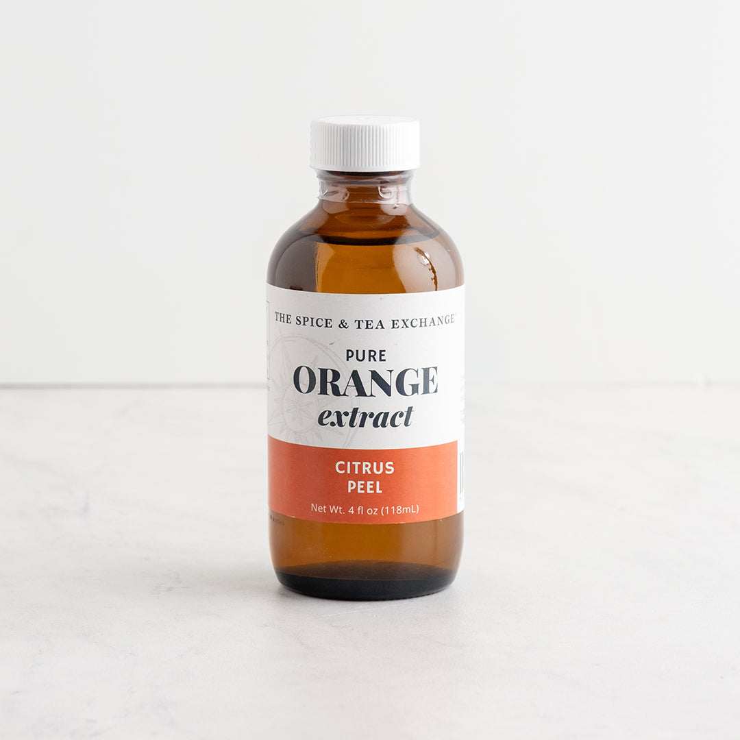 An amber glass bottle of Pure Orange Extract by The Spice & Tea Exchange, featuring a white cap and a white label with a coral orange-red band at the bottom, set against a clean white background.