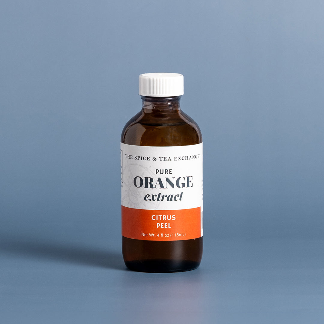 A bottle of Tree Spice & Tea Exchange Pure Orange Extract (Citrus Peel) in an amber glass bottle with a white cap and white label featuring an orange accent band, set against a solid blue-gray background.