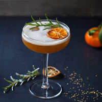 A frothy amber cocktail in a coupe glass, garnished with a rosemary sprig and a dried orange slice, set on a dark surface with scattered seeds and a blurred orange in the background.