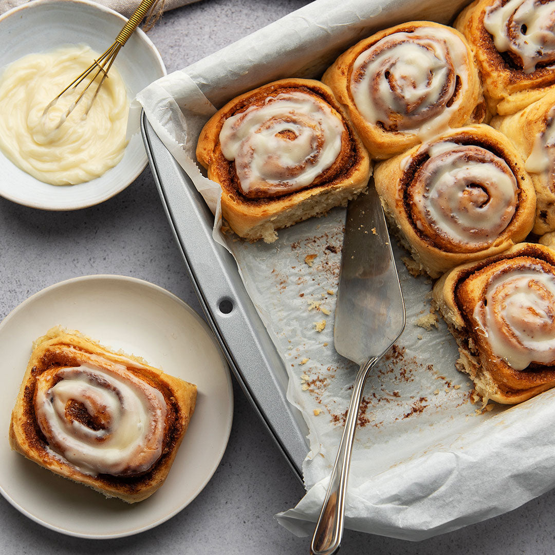 Cinnamon rolls with white icing on a baking sheet, served on small plates with a bowl of cream frosting and a whisk nearby.