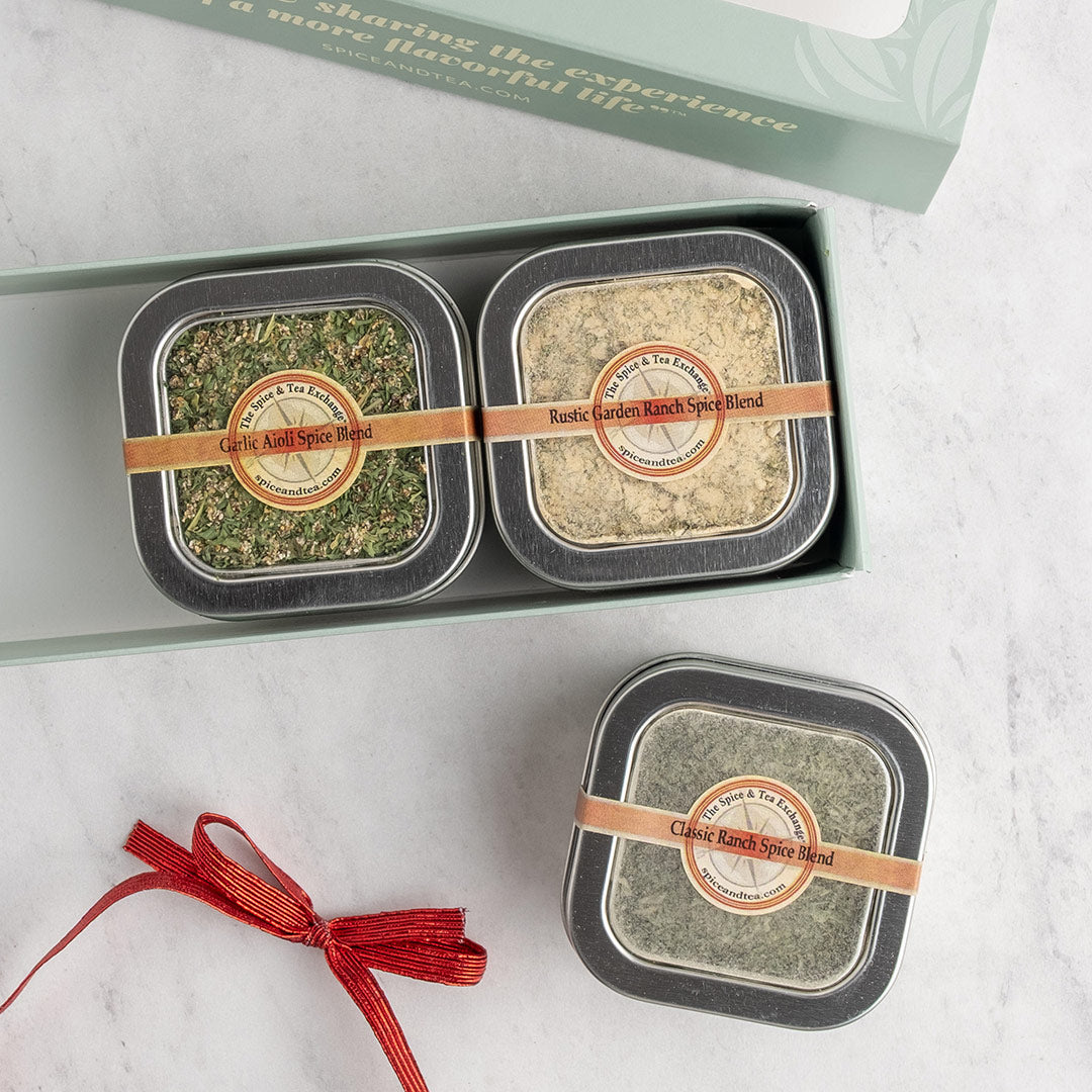 A gift set of three square metal tins of spice blends arranged in an open pale green box on a light marble surface, with one tin placed below and a red ribbon setting next to it. The tins have round labels.