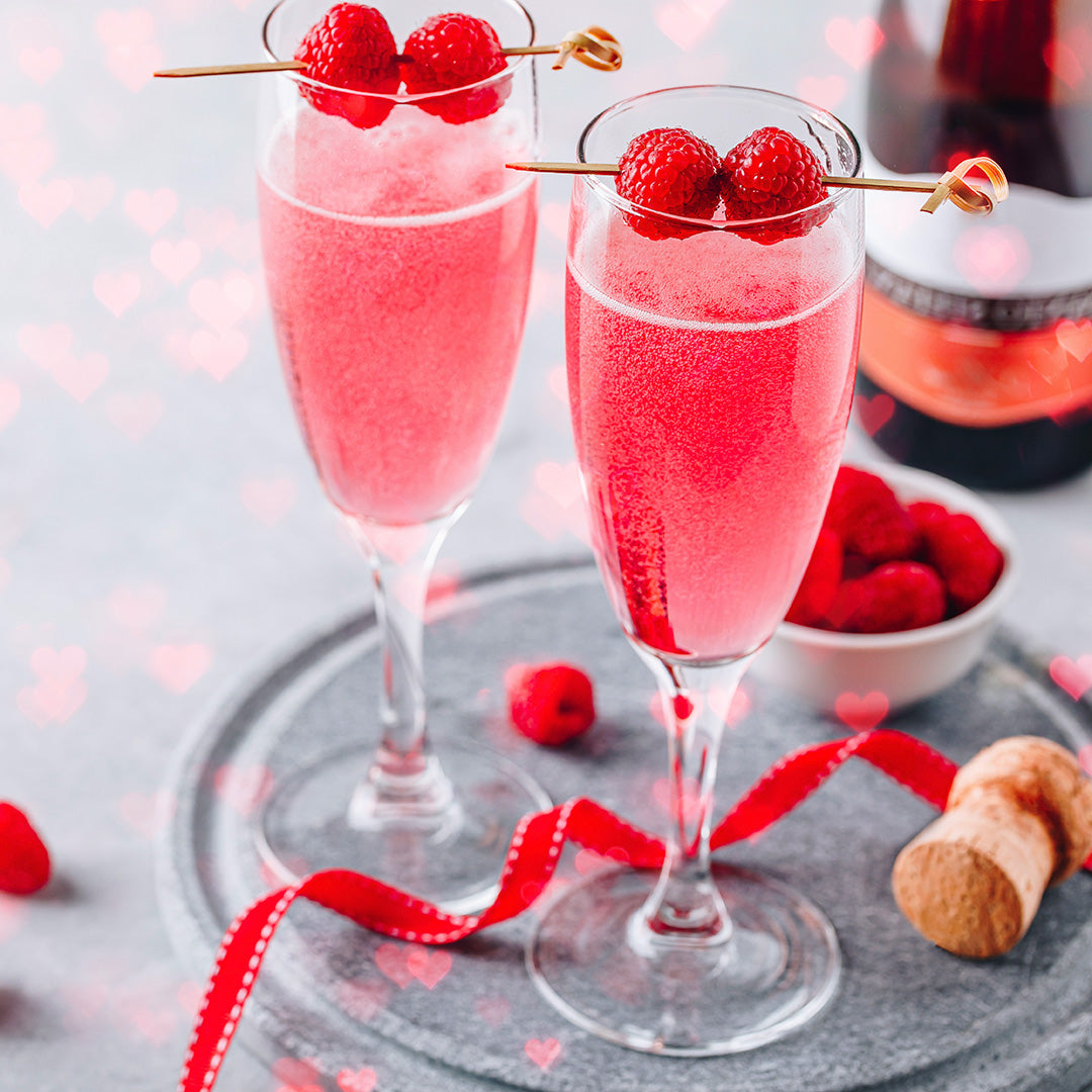 Two pink drinks in champagne flutes with raspberries on a gray surface.
