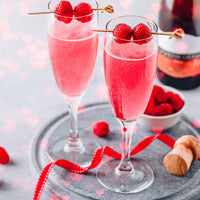 Two pink drinks in champagne flutes with raspberries on a gray surface.
