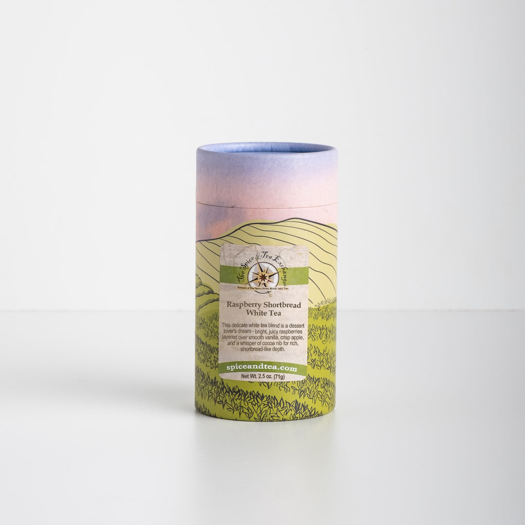 A cylindrical tea container features a pastel sky and landscape design. The label reads Raspberry Shortbread White Tea and highlights notes of raspberry, white chocolate, product details, and a website URL.