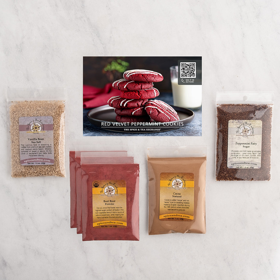 A cookie recipe and ingredient kit arranged on a light marble surface, including a recipe card showing red velvet peppermint cookies and several bags of kit contents/baking ingredients.
