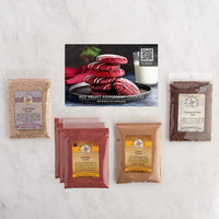 A cookie recipe and ingredient kit arranged on a light marble surface, including a recipe card showing red velvet peppermint cookies and several bags of kit contents/baking ingredients.