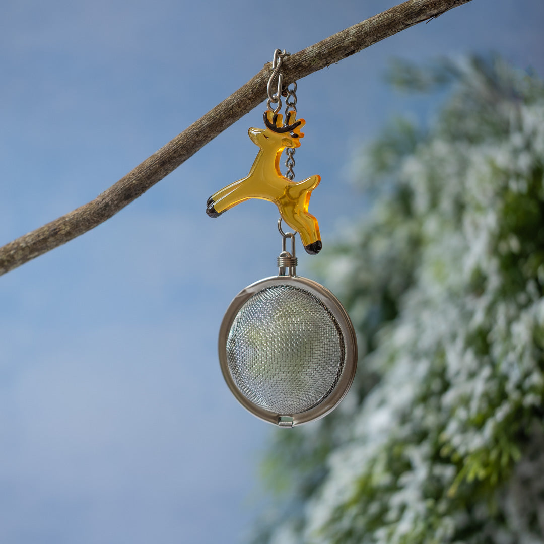 Tea infuser with reindeer design hanging from a branch against a blue sky and green foliage background