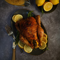 A roasted whole chicken on a dark oval platter, garnished with lemon slices, rosemary, and herbs, set on a gray stone-like surface with a fork, knife, and additional lemons nearby.