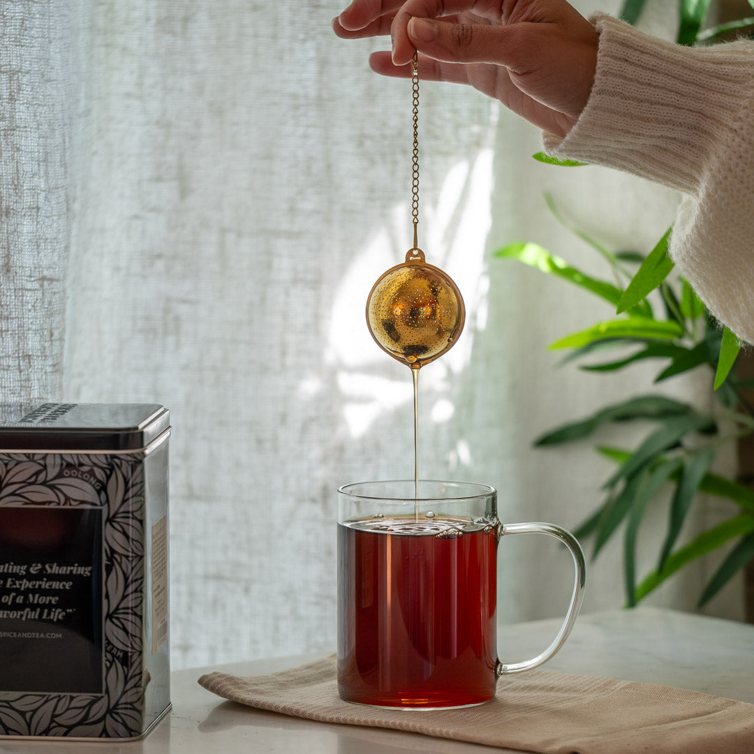 Tea bag being added to a cup of tea with a decorative tea ball, on a light surface with a plant in the background.