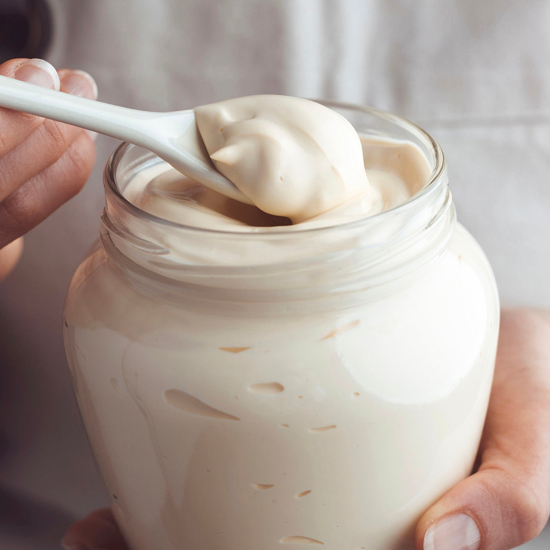 A close-up of a hand holding a spoonful of thick, creamy white mayonnaise above an open jar, next to a jar of Roasted Espresso Monk Fruit Sweetener.