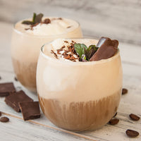 Two glass cups of creamy iced coffee made with Roasted Espresso Monk Fruit Sweetener are topped with whipped cream, chocolate shavings, fresh mint, and chocolate pieces. Coffee beans and chocolate squares decorate a light wooden surface.
