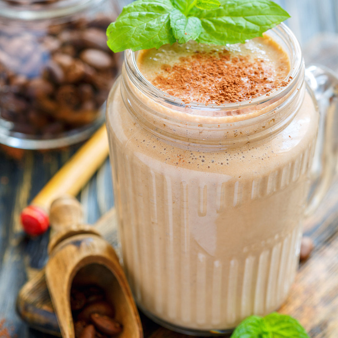 A creamy iced coffee smoothie in a glass jar, topped with cocoa powder and mint, sits beside coffee beans, a wooden scoop, and a jar of Roasted Espresso Monk Fruit Sweetener on a rustic wooden surface.